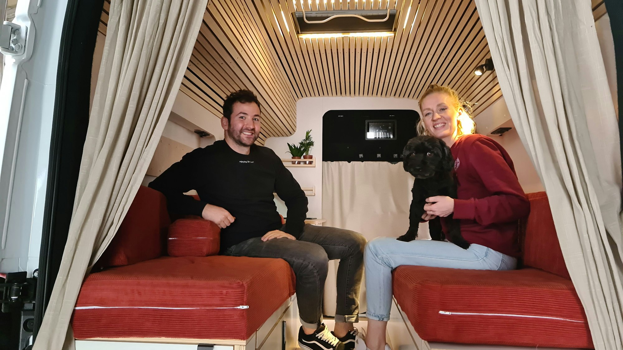 Ein junger Mann mit dunklem Haar und eine junge blonde Frau sitzen sich in einem Transporter auf Polsterbänken gegenüber. Die Frau hat einen schwarzen Hund auf dem Schoß.