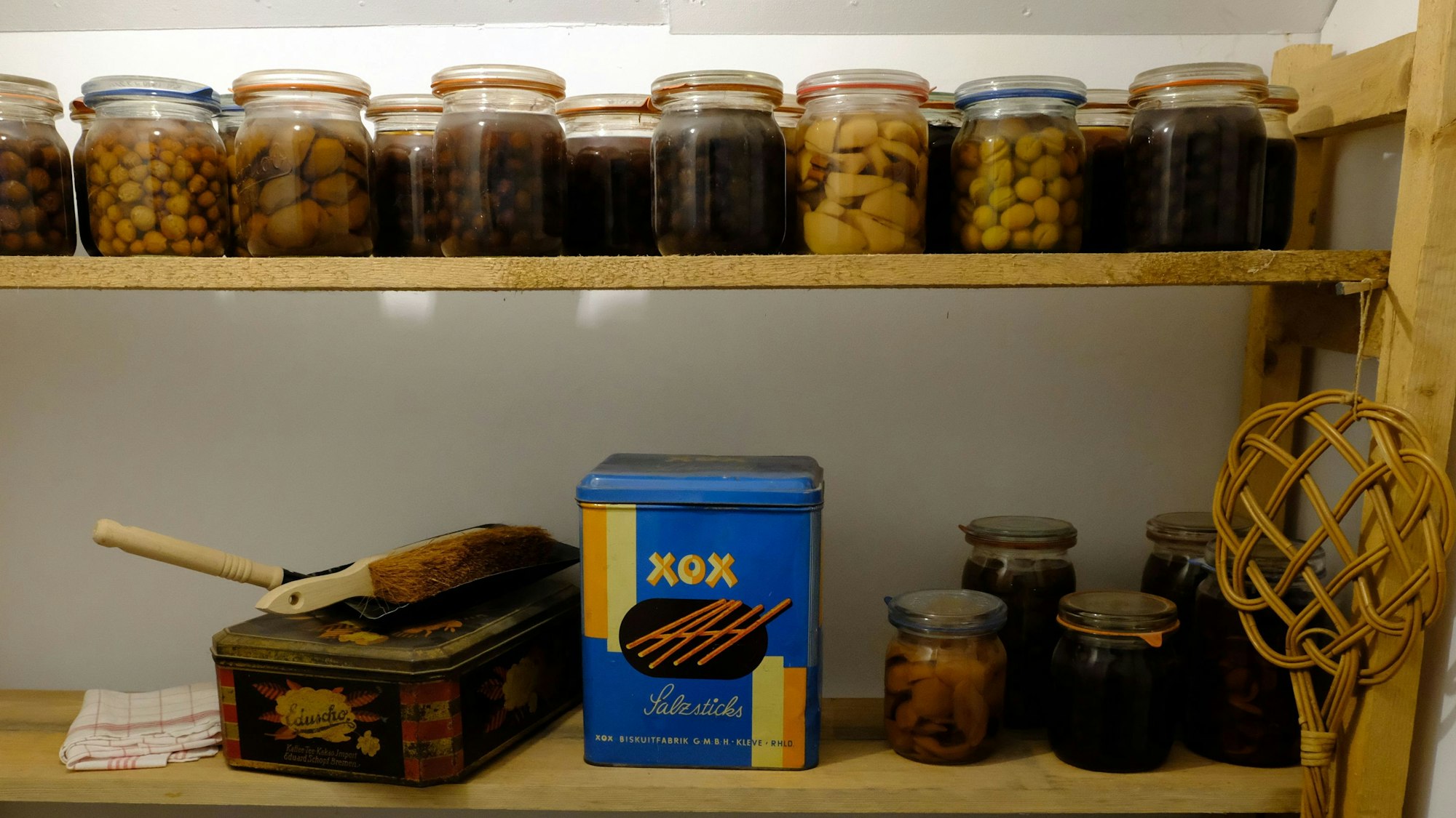 Auf einem Holzregal stehen verschieden gefüllte Einmachgläser. Außerdem sind ein Teppichklopfer, eine Kehrgarnitur und eine blaue Metalldose zu sehen.