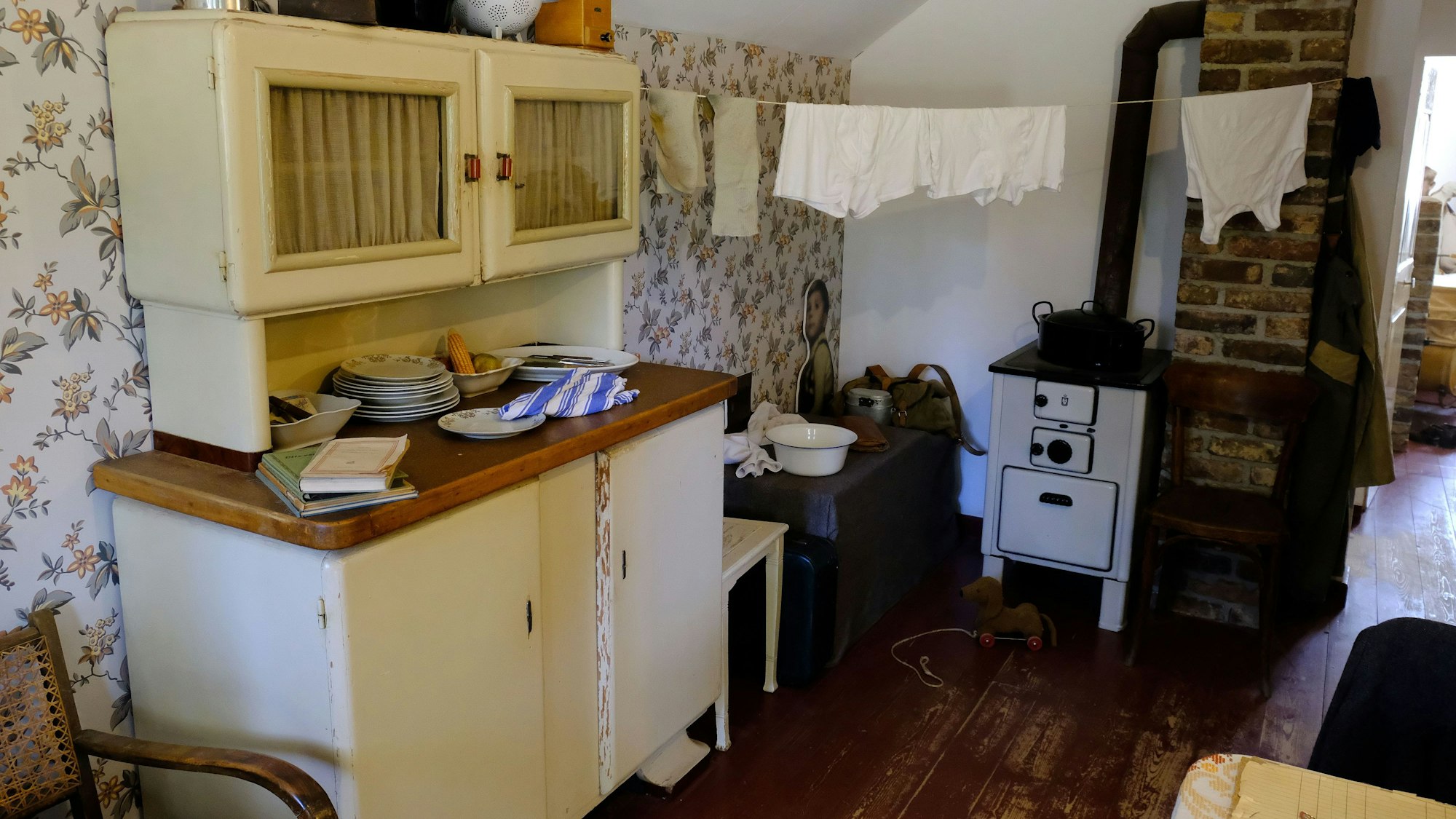 In einem Innenraum der Nissenhütte steht ein cremeweißer Buffetschrank, über dem Kohleofen hängen Wäschestücke zum Trocknen auf einer Leine.