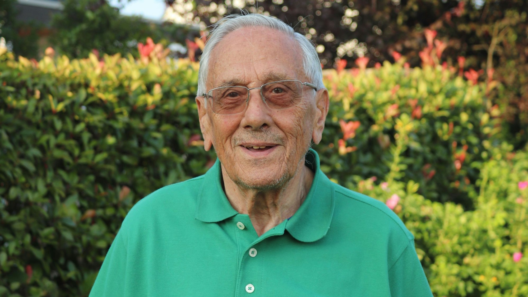 Ein Mann im grünen Polohemd steht vor einer Hecke in einem Garten.