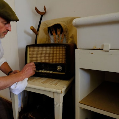 Peter Drespa verkörpert einen Kriegsheimkehrer des Zweiten Weltkriegs im Freilichtmuseum Kommern. In der Kleidung der späten 1940-er Jahre kniet er vor einem Radio, das auf einem Küchenstuhl steht.