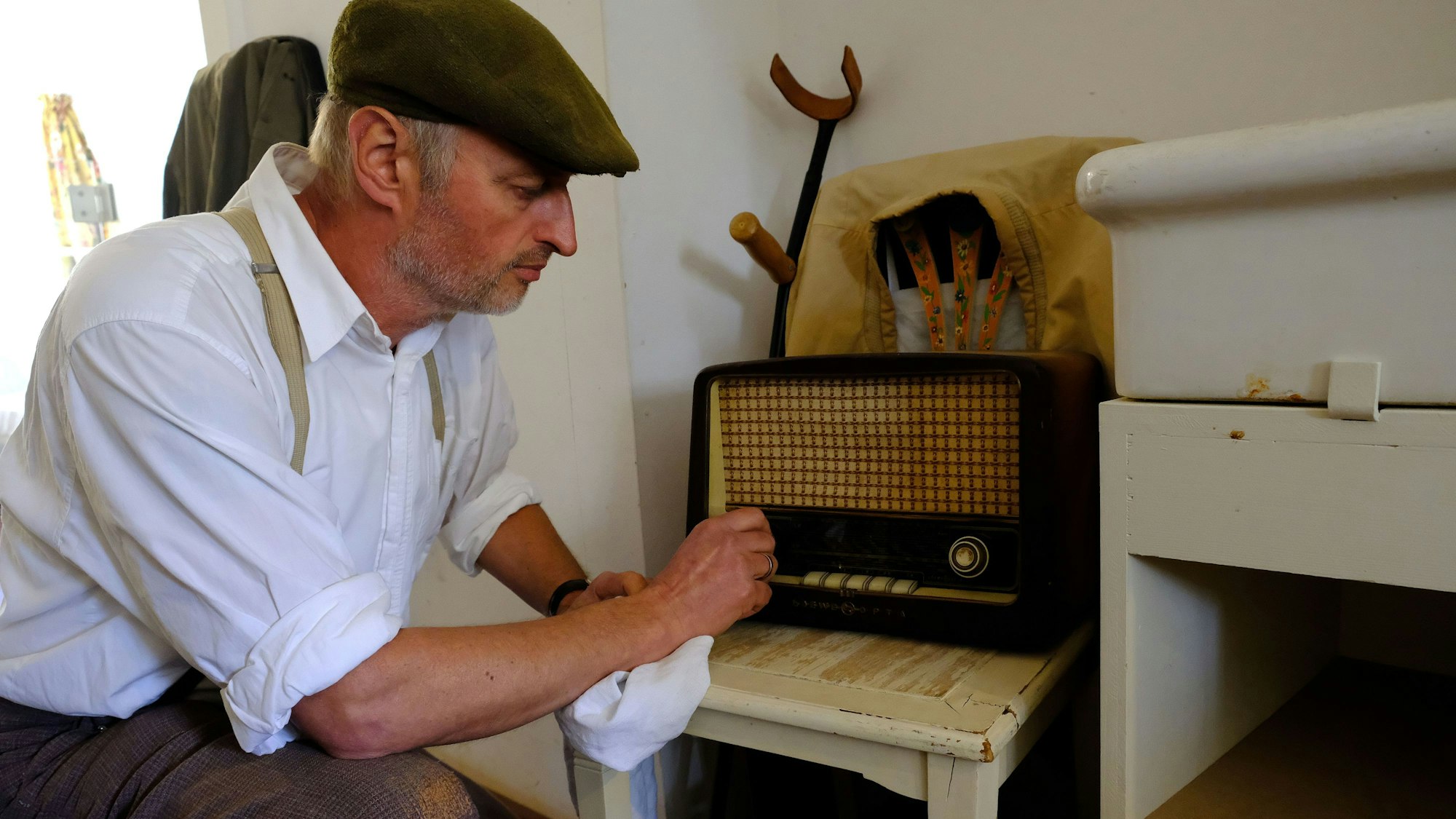 Peter Drespa verkörpert einen Kriegsheimkehrer des Zweiten Weltkriegs im Freilichtmuseum Kommern. In der Kleidung der späten 1940-er Jahre kniet er vor einem Radio, das auf einem Küchenstuhl steht.