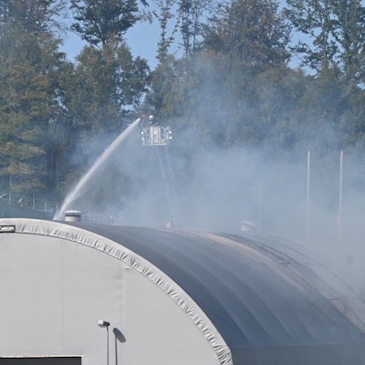 Ein Feuerwehrmann löscht von einer Drehleiter aus eine brennende Halle mit Zeltdach.