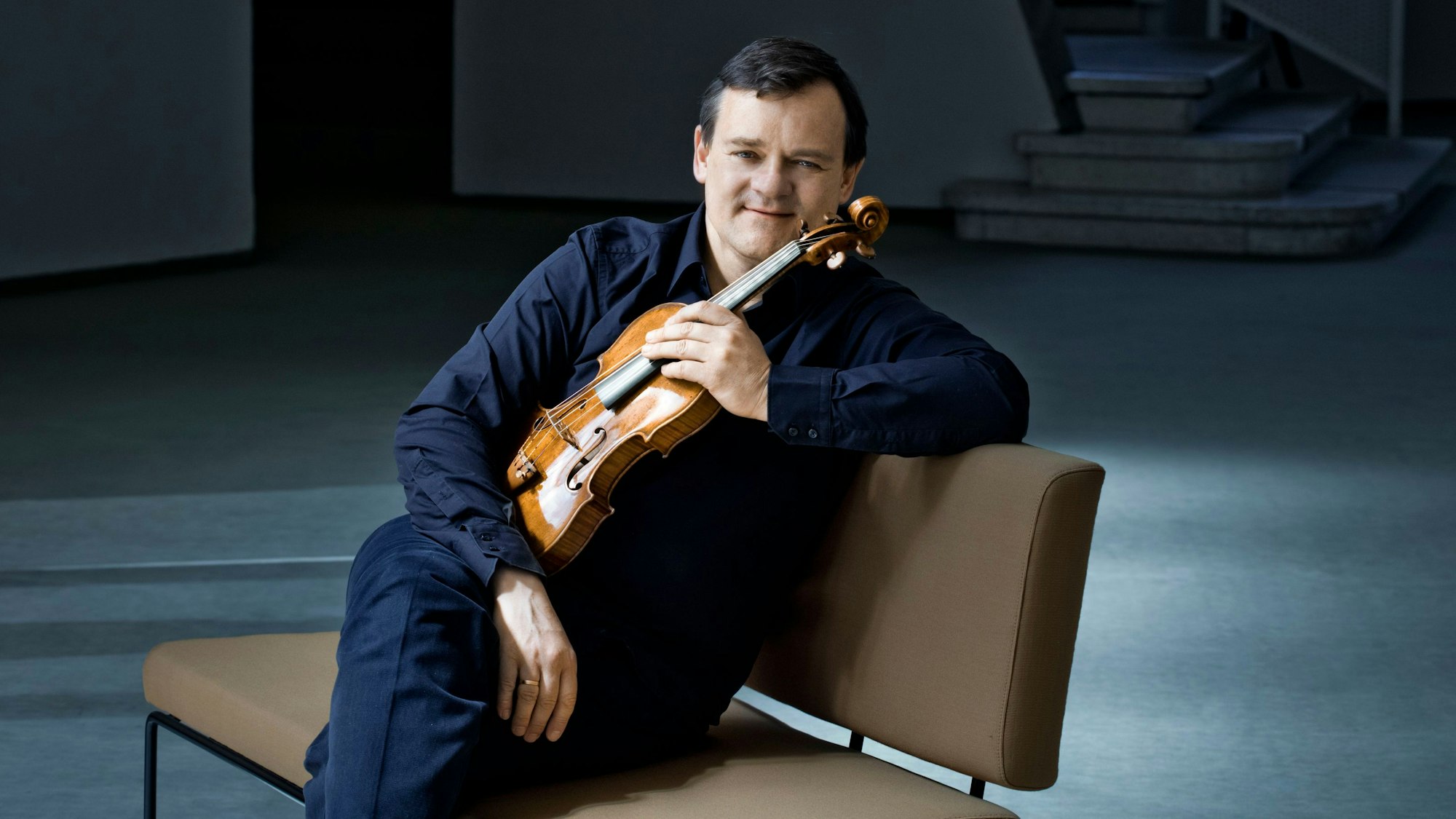 Frank Peter Zimmermann sitzt mit Geige auf einem Stuhl.