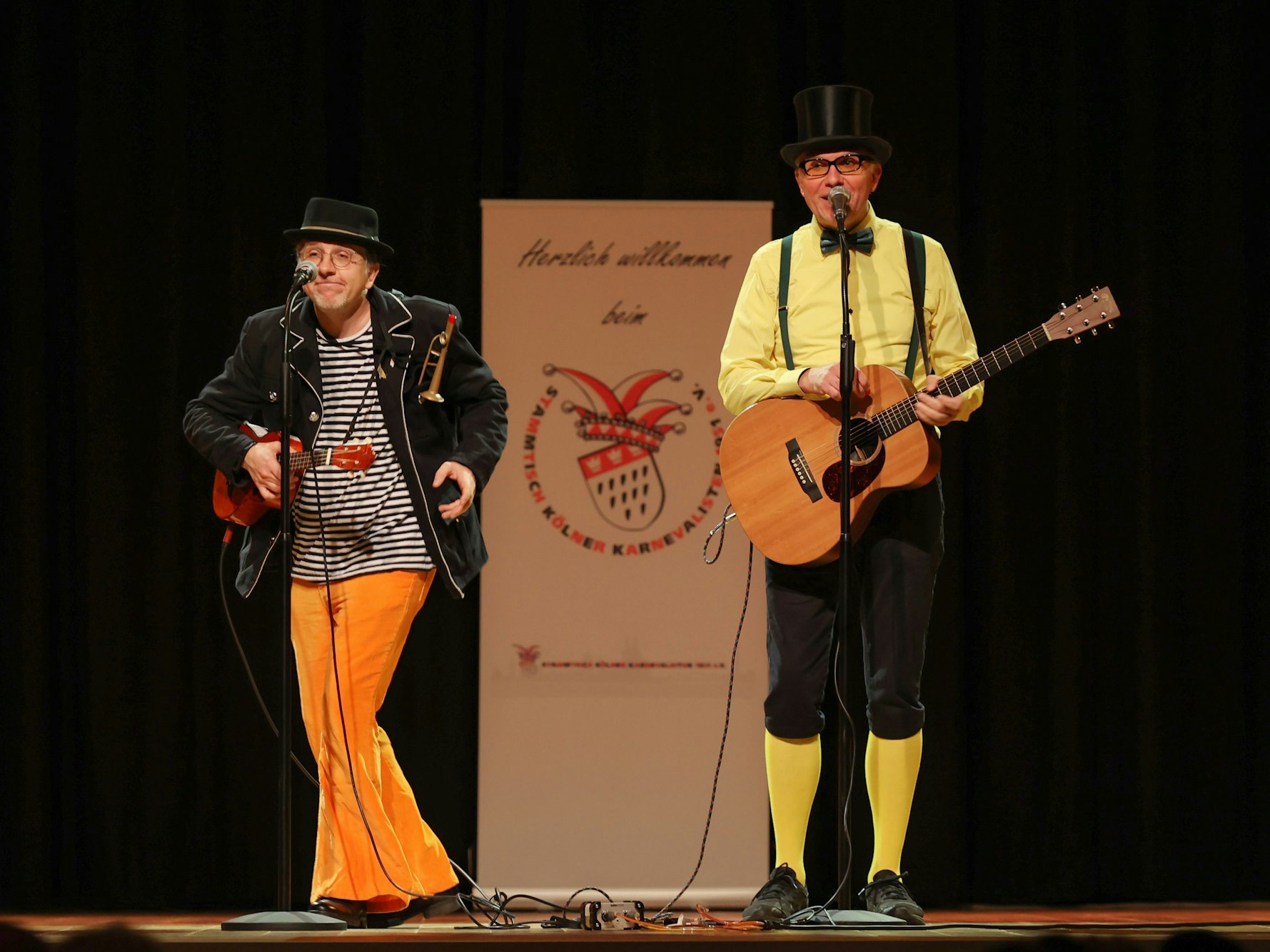 Zwei Männer stehen mit Gitarre, Hüten und bunten Klamotten auf der Bühne.