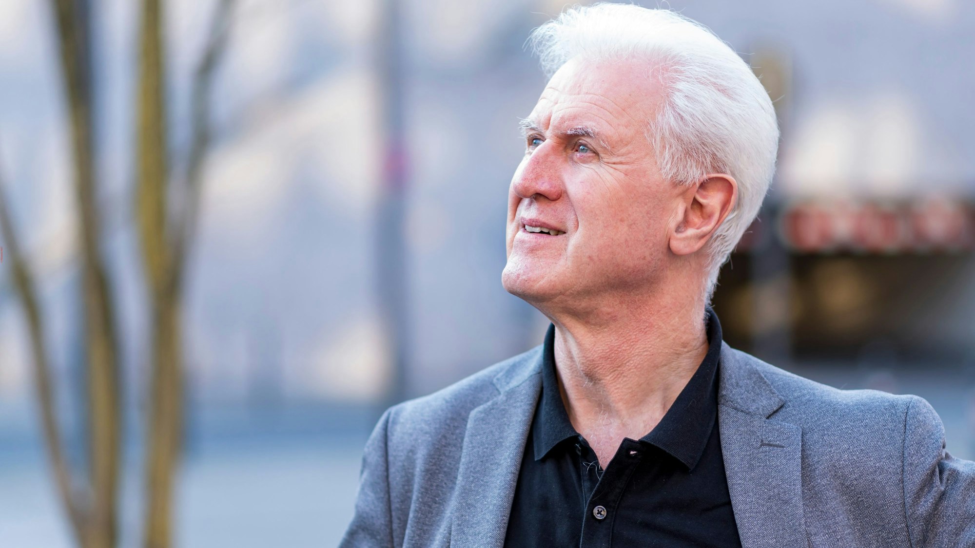 Christoph Poppen trägt ein blaugraues Sakko zum schwarzen Hemd und schaut nach oben.