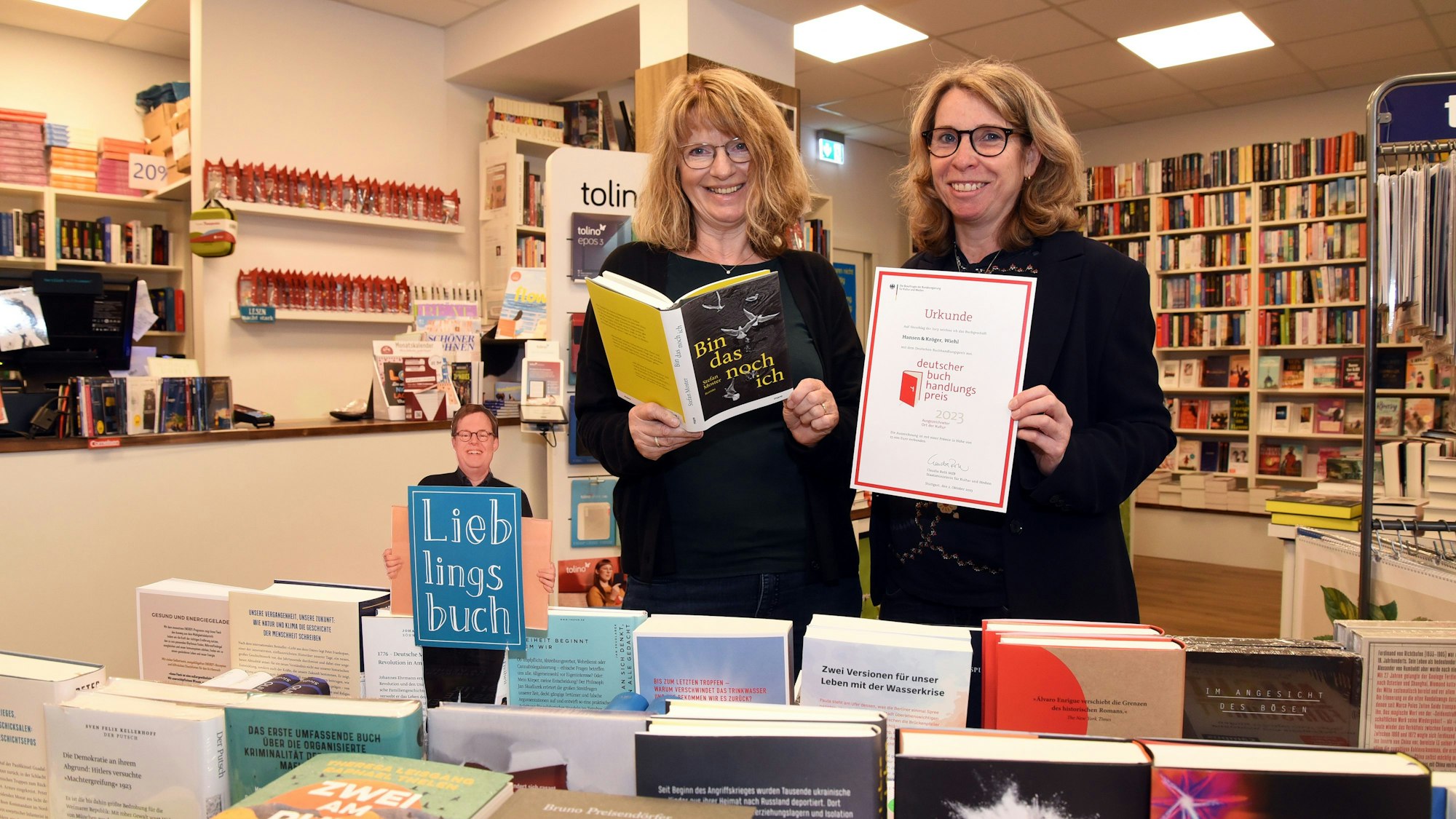 Zwei Frauen stehen in einer Buchhandlung. Eine hält ein Buch in der Hand. Die andere eine Urkunde.