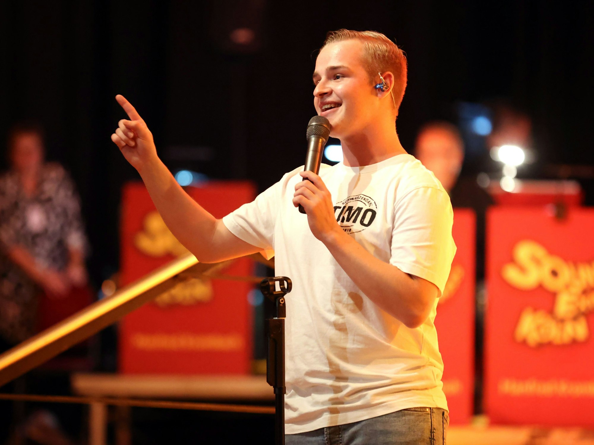 Timo Schwarzendahl in weißem T-Shirt und Mikrofon auf der Bühne.