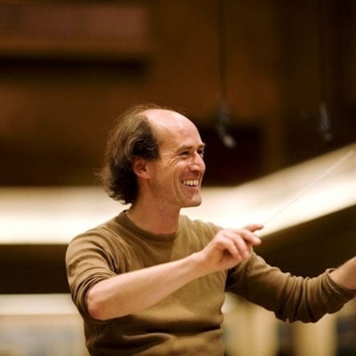 Peter Rundel hat den rechten Ärmel seines braunen Pullis hochgekrämpelt, er lächelt und hat die Hände zum Dirigat vor sich erhoben.