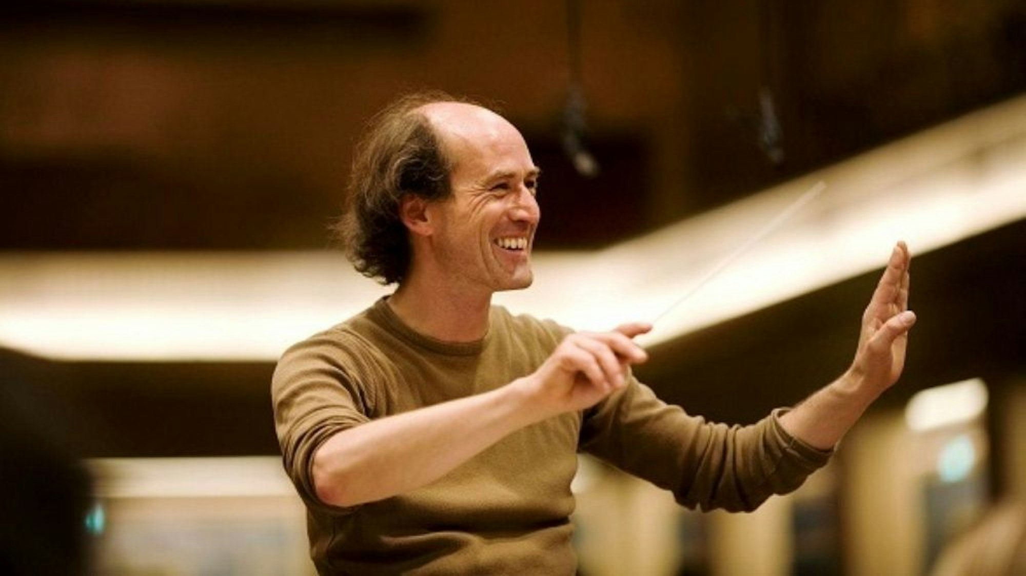 Peter Rundel hat den rechten Ärmel seines braunen Pullis hochgekrämpelt, er lächelt und hat die Hände zum Dirigat vor sich erhoben.