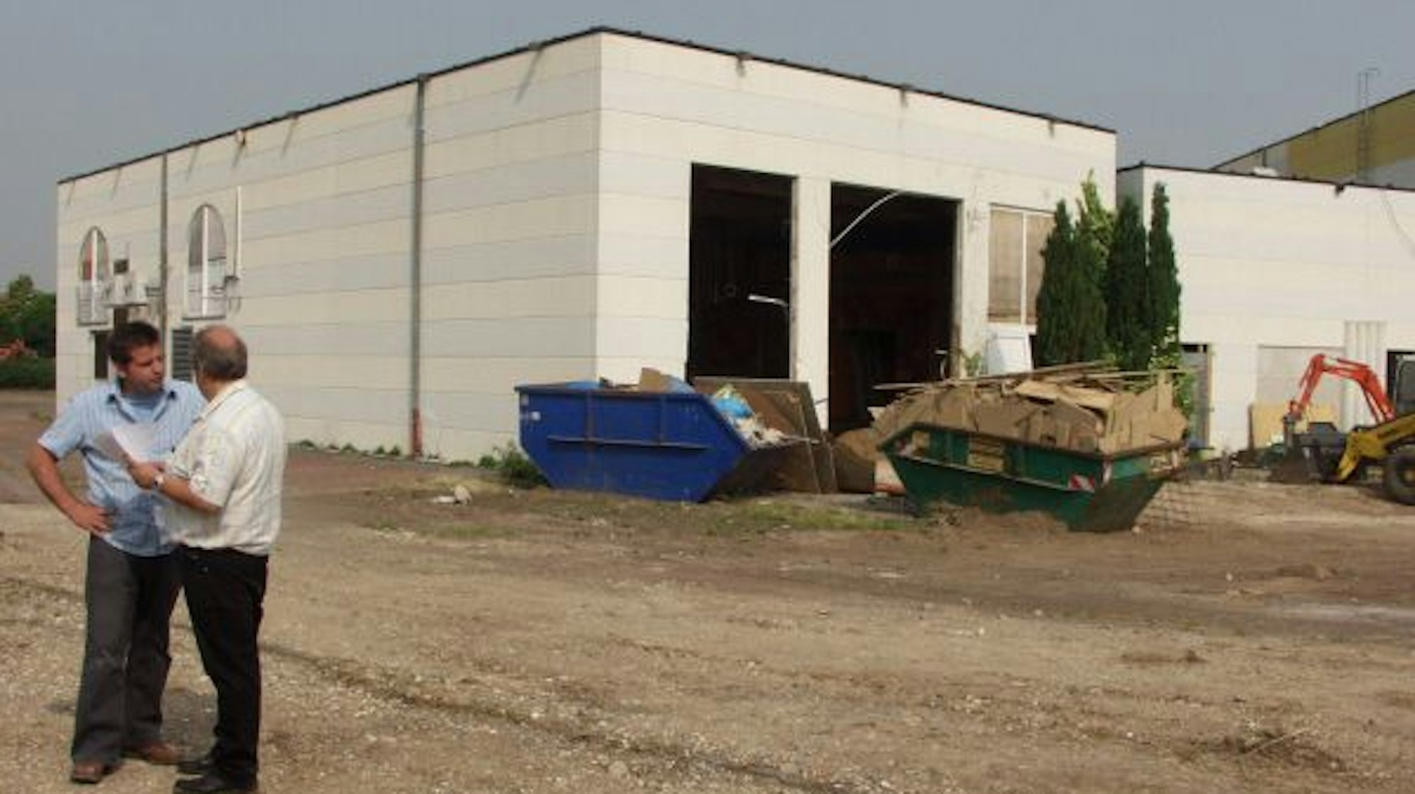 Links im Bild unterhalten sich zwei Männer. Im Mittelpunkt steht eine große Halle, davor Container.