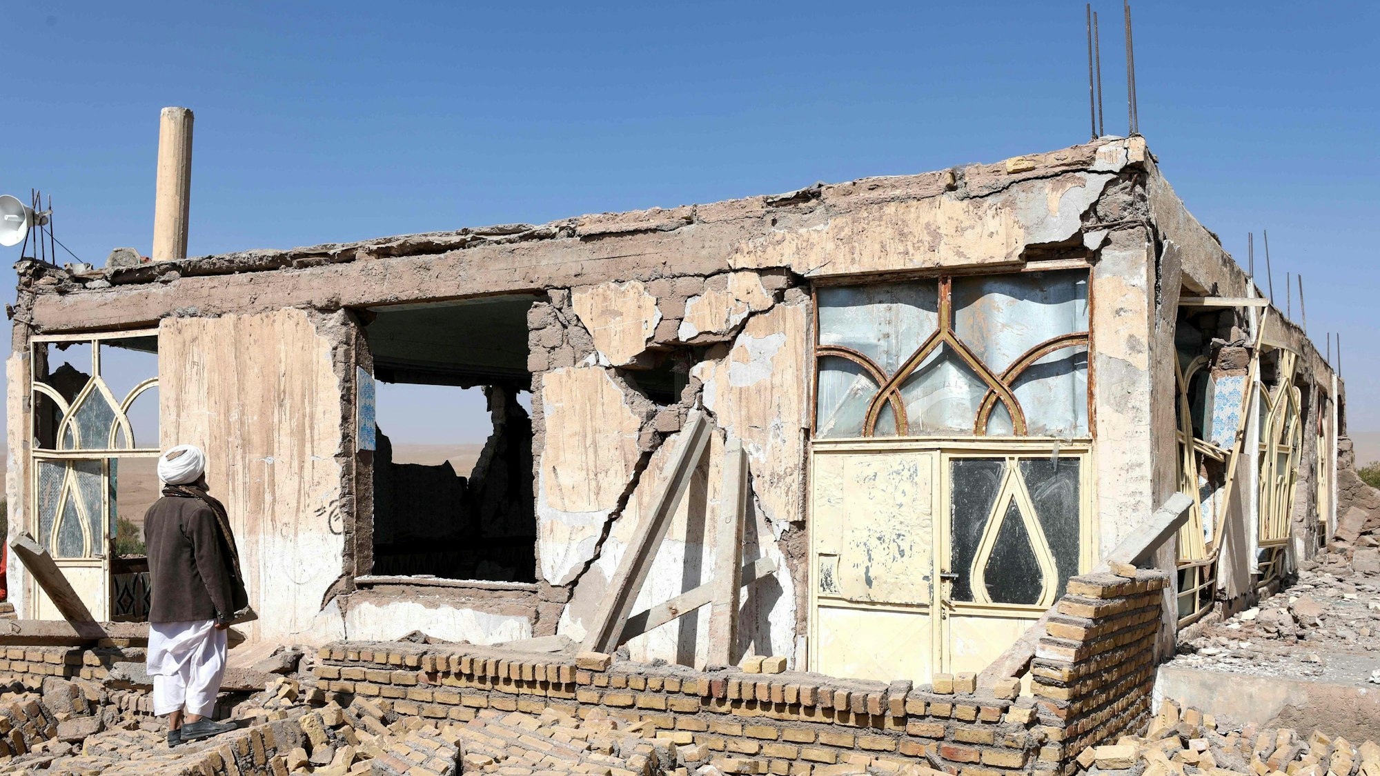 Ein Mann mit Turban steht vor einem zerstörten Haus unter einem wolkenlosen Himmel.