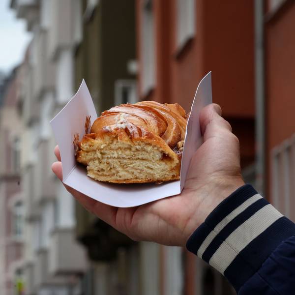 Eine Hand hält eine Zimtschnecke hoch, im Hintergrund der Colonius und ein Kölner Straßenzug.