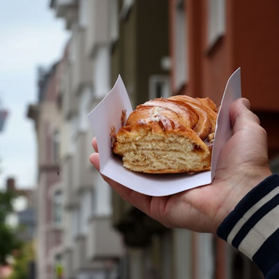 Eine Hand hält eine Zimtschnecke hoch, im Hintergrund der Colonius und ein Kölner Straßenzug.