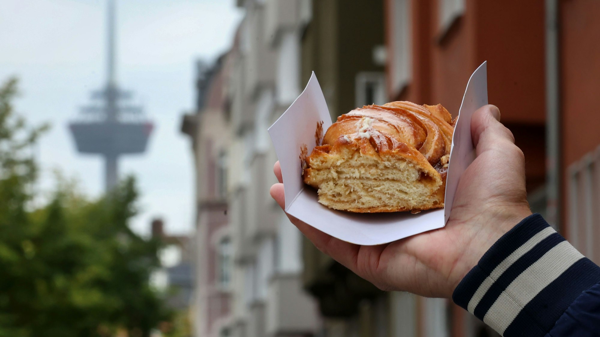 Eine Hand hält eine Zimtschnecke hoch, im Hintergrund der Colonius und ein Kölner Straßenzug.