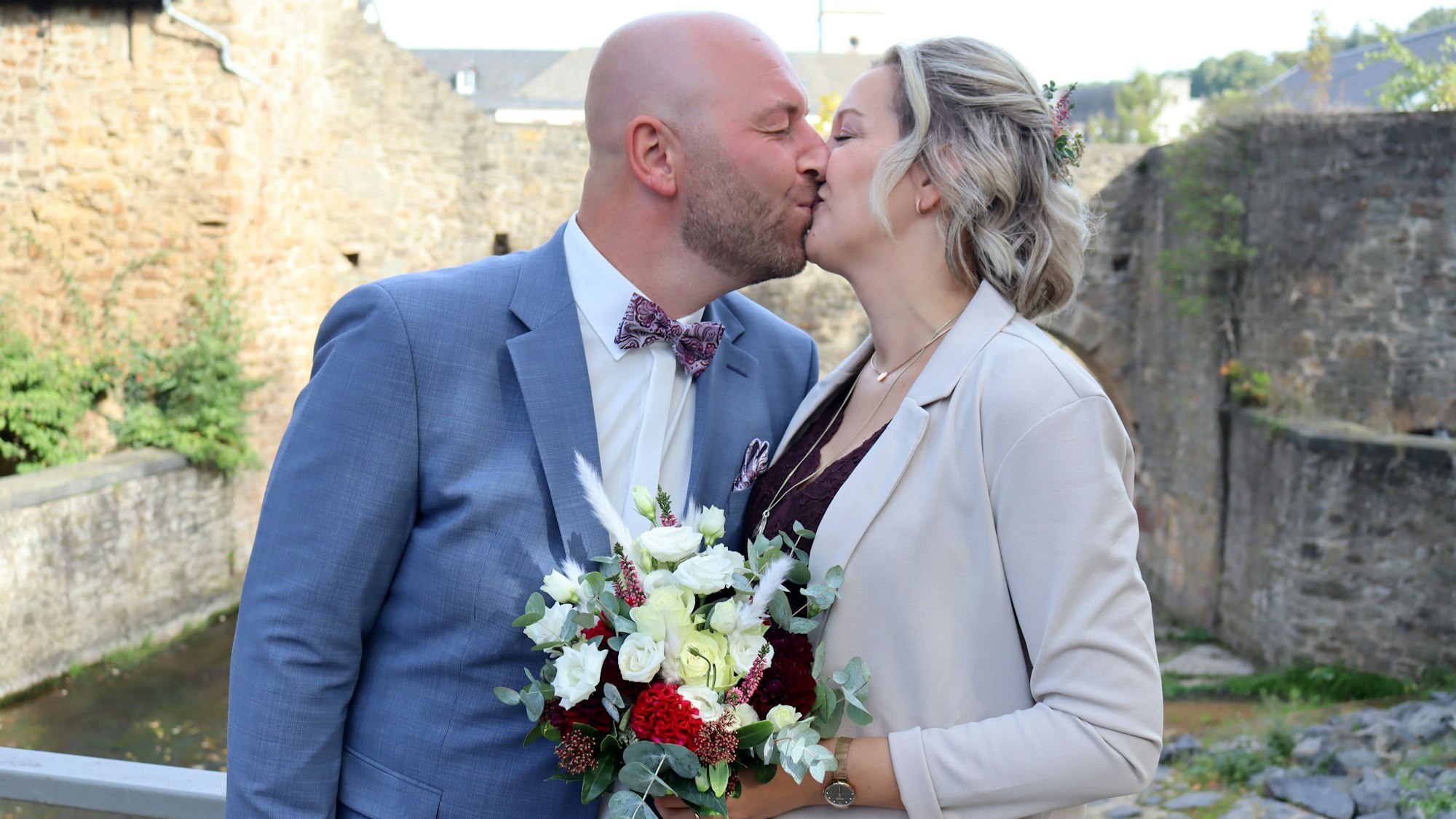 Das frisch verheiratete Ehepaar steht auf einer Brücke in Bad Münstereifel und küsst sich.