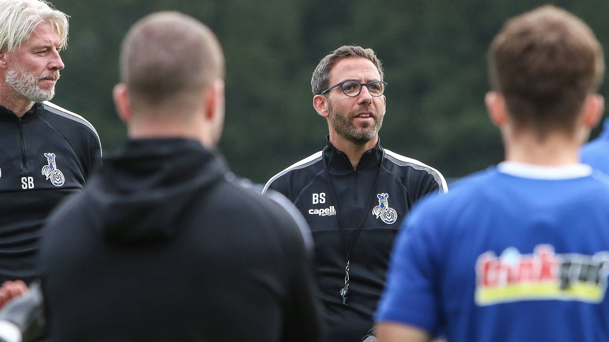 Trainer Boris Schommers spricht zu den Duisburger Fußballern, die im Halbkreis vor ihm stehen.