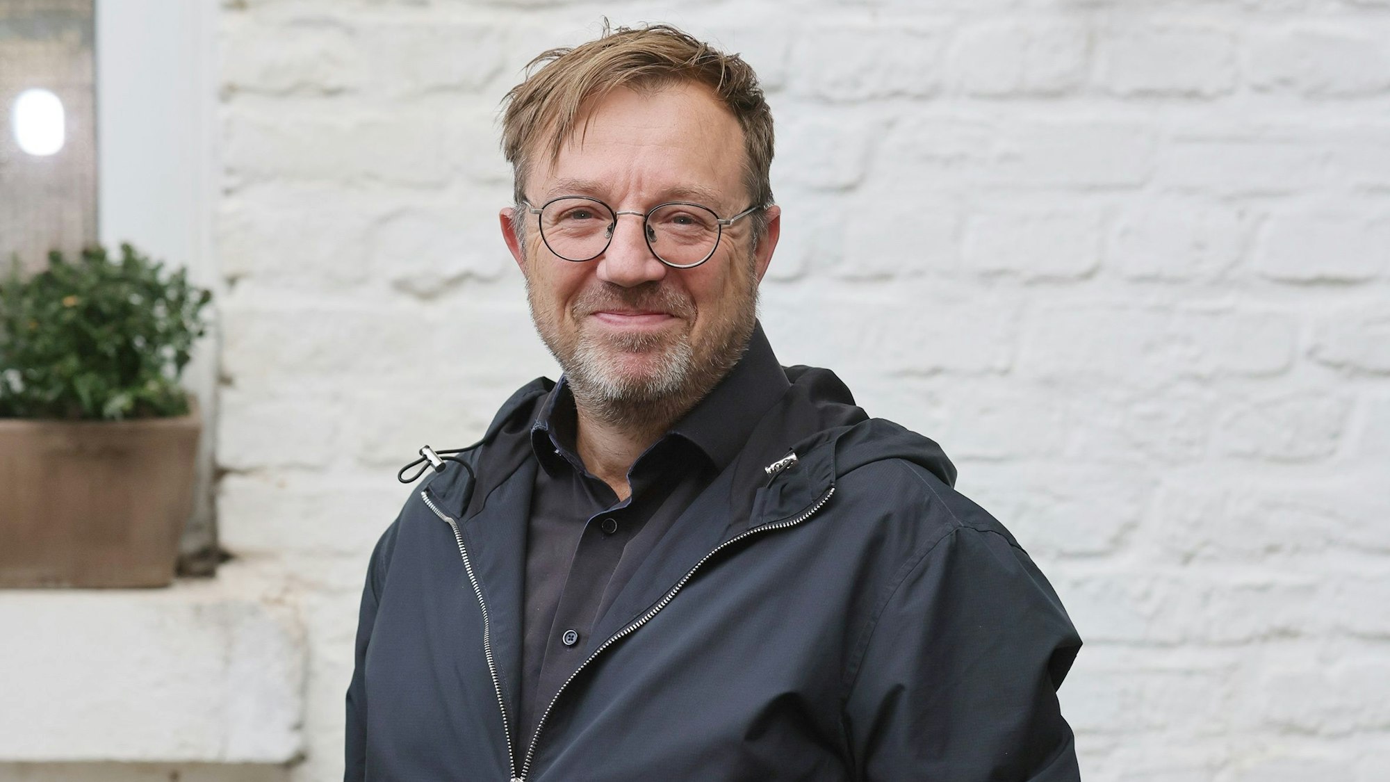 Holger Recktenwald lächelt in die Kamera. Er steht vor einer weißen Wand, trägt eine schwarze Jacke und eine Brille.