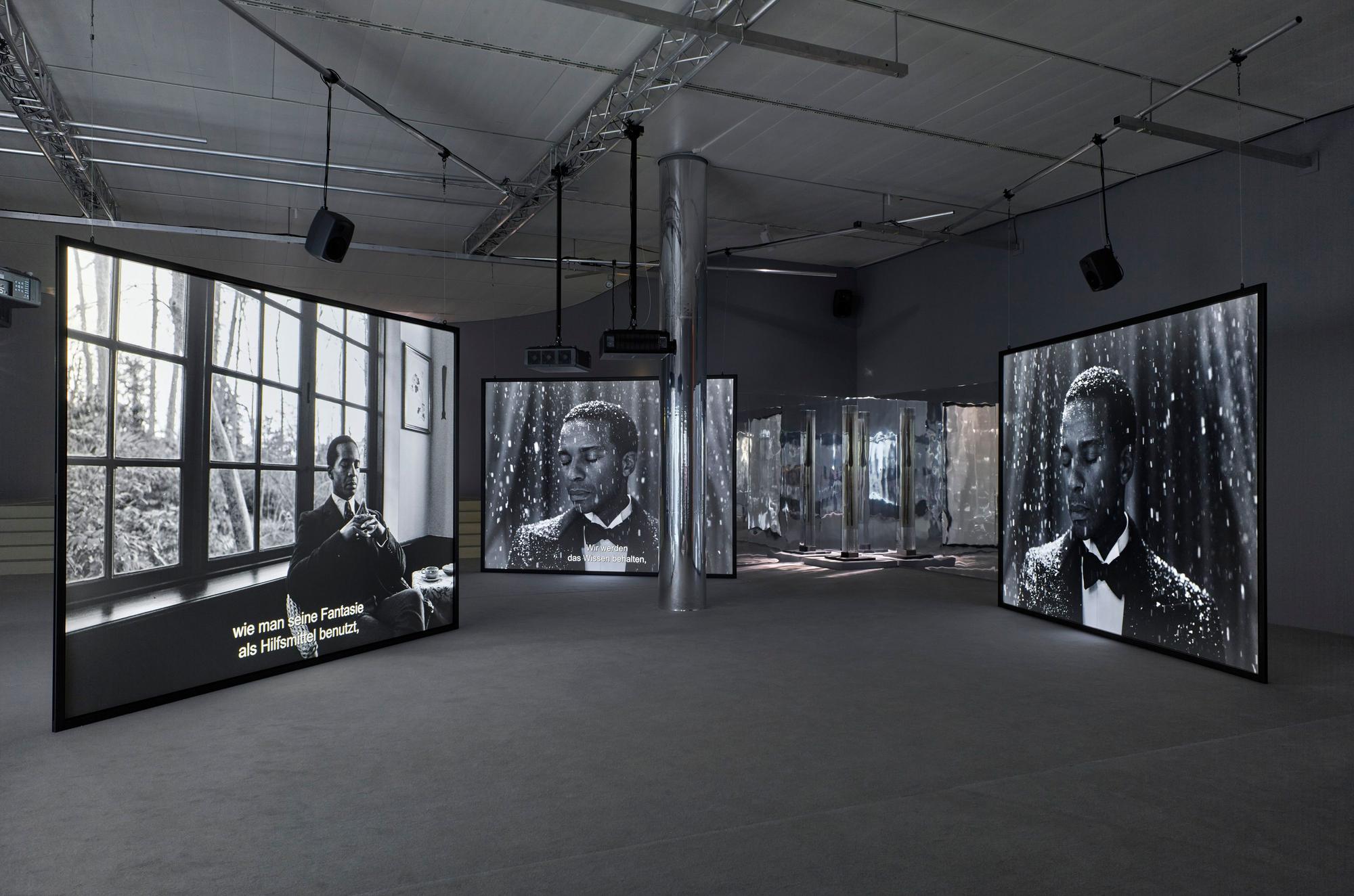 Auf dem schwarz-weiß Foto ist die Installation zu sehen. Mehrere große Leinwände zeigen Fotografien.