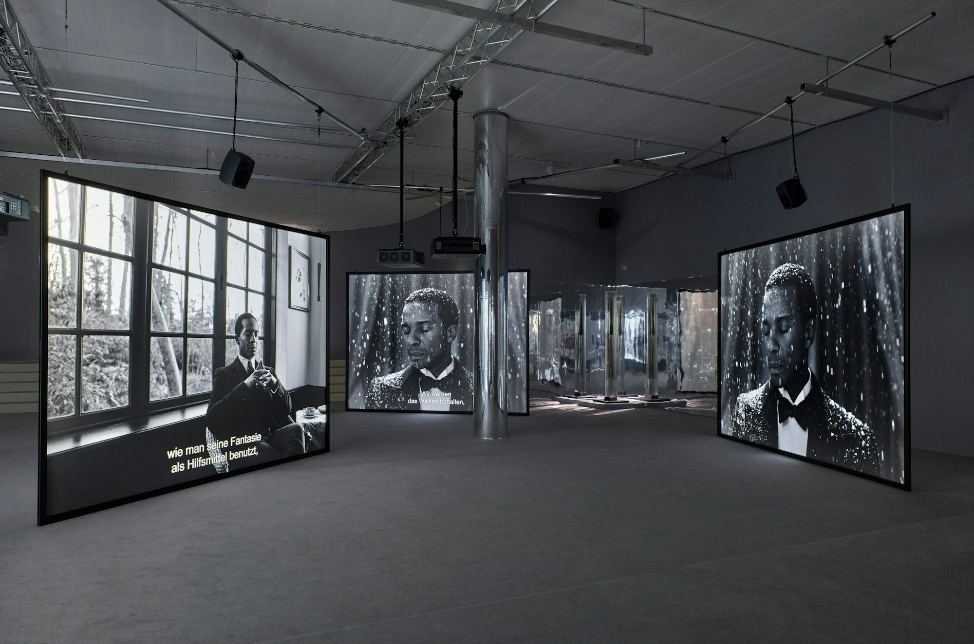 Auf dem schwarz-weiß Foto ist die Installation zu sehen. Mehrere große Leinwände zeigen Fotografien.
