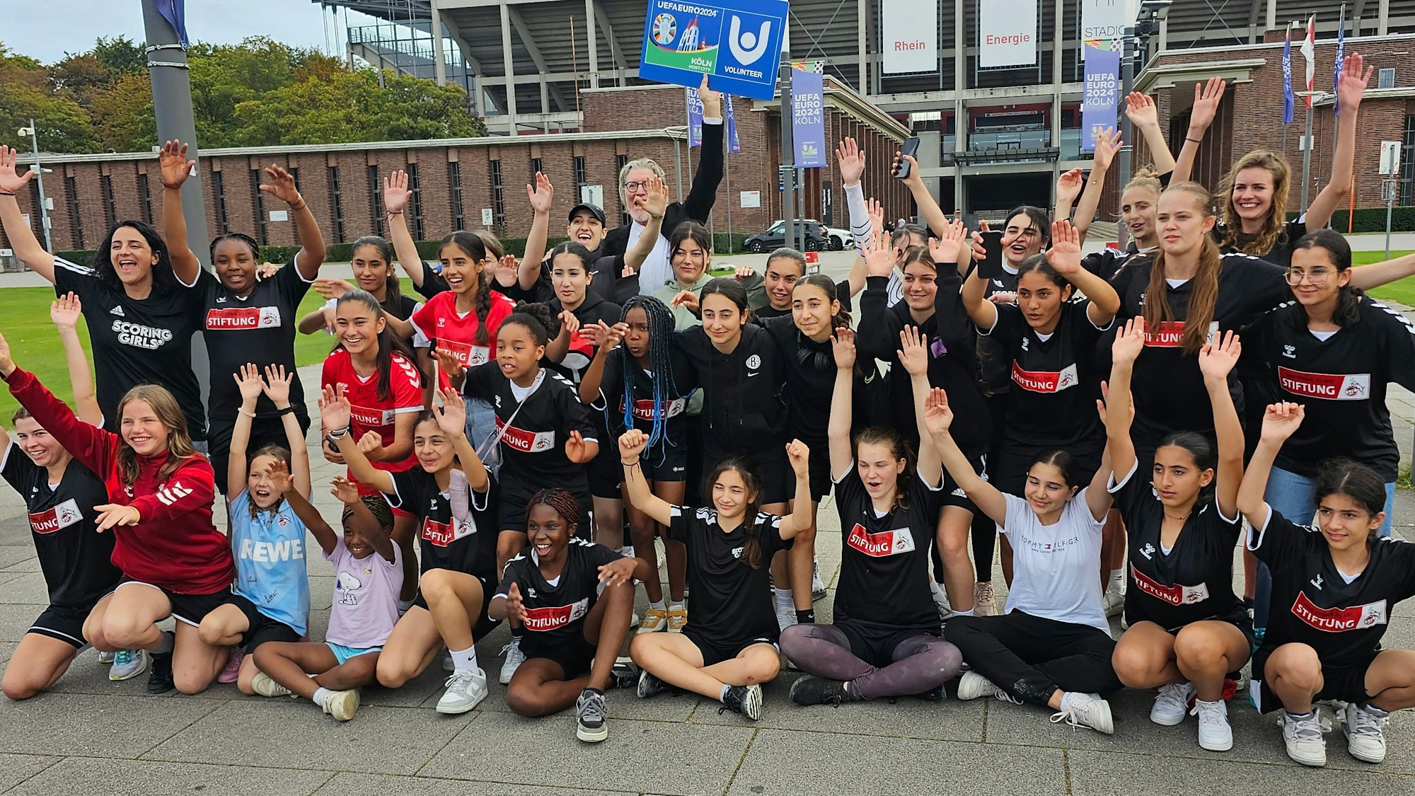 Gruppe von Mädchen im Fußballtrikot wirft vor einem Fußballstadion die Arme hoch.