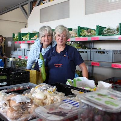 Drei Mitarbeiterinnen der Tafel laufen an den Kisten mit Obst und Gemüse vorbei, um die Taschen der Kunden zu füllen.