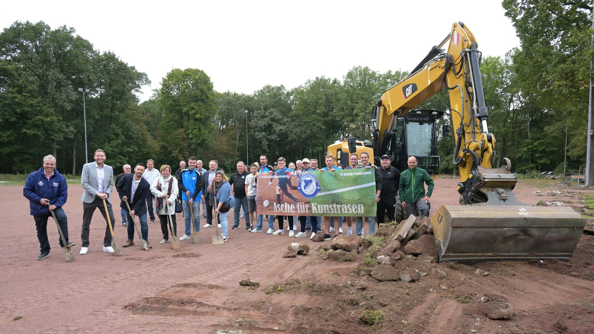 Vereinsmitglieder, darunter Vorstandsmitglieder und stellvertretende Bürgermeisterin, stehen auf dem roten Aschenplatz neben dem Abbruchbagger.