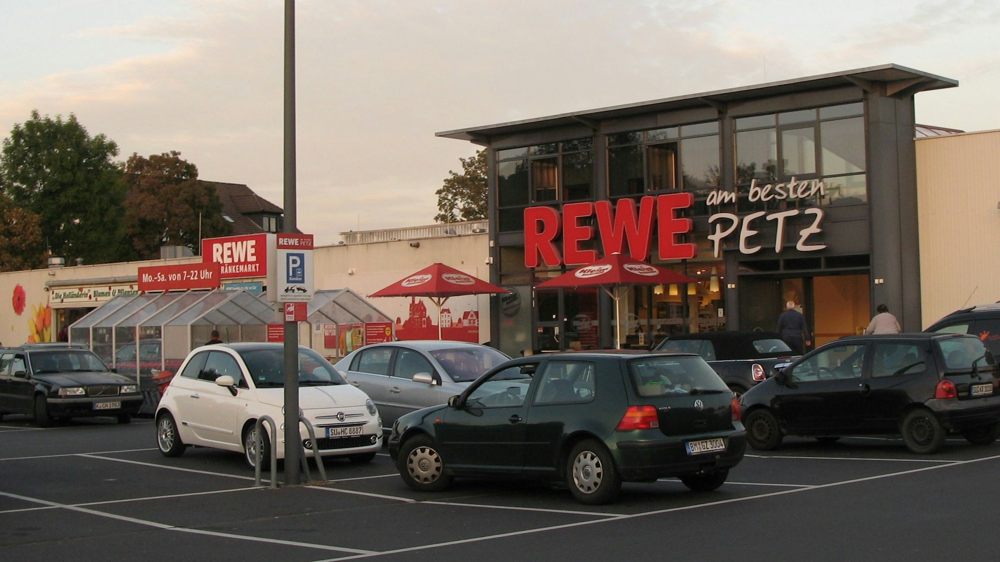 Das Bild zeigt die Front des Rewe-Supermarkts mit davor stehenden Autos von Kunden.