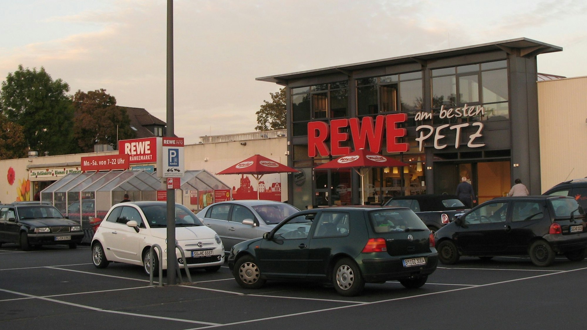 Das Bild zeigt die Front des Rewe-Supermarkts mit davor stehenden Autos von Kunden.
