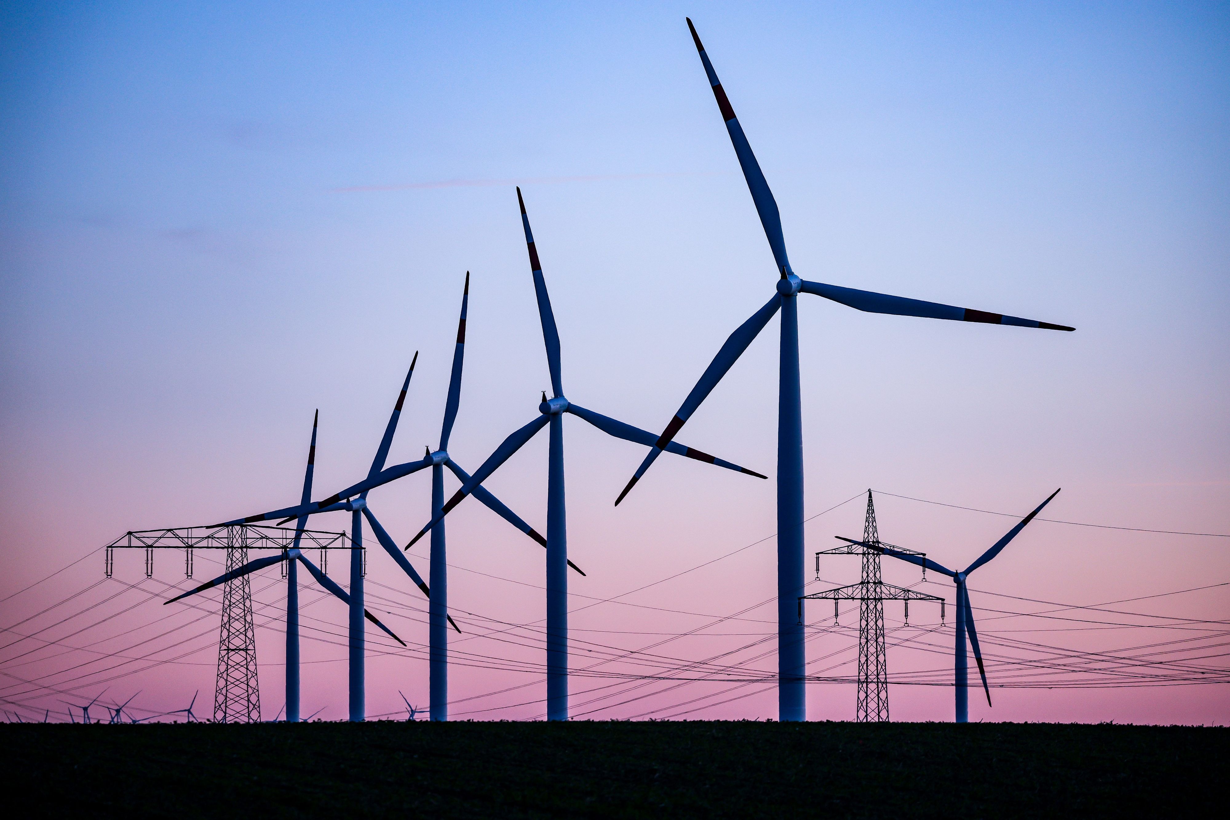 Windkraftanlagen und Hochspannungsleitungen sind vor dem Abendhimmel bei Leuna zu sehen. Der Ausbau von Windrädern an Land kommt in Deutschland  voran.