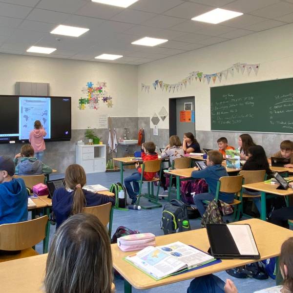Ein Schülerin steht neben dem Lehrer an der digitalen Tafel. Die anderen sitzen auf ihren Plätzen und haben Tabletcomputer vor sich.