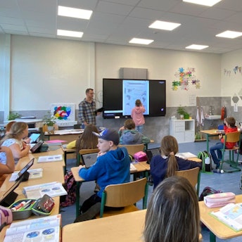 Ein Schülerin steht neben dem Lehrer an der digitalen Tafel. Die anderen sitzen auf ihren Plätzen und haben Tabletcomputer vor sich.
