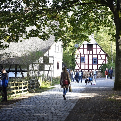 Mehrere Menschen laufen an Fachwerkhäusern im Freilichtmuseum vorbei. Ein hoher Baum rechts im Bild wirft Schatten auf die gepflasterten Wege.