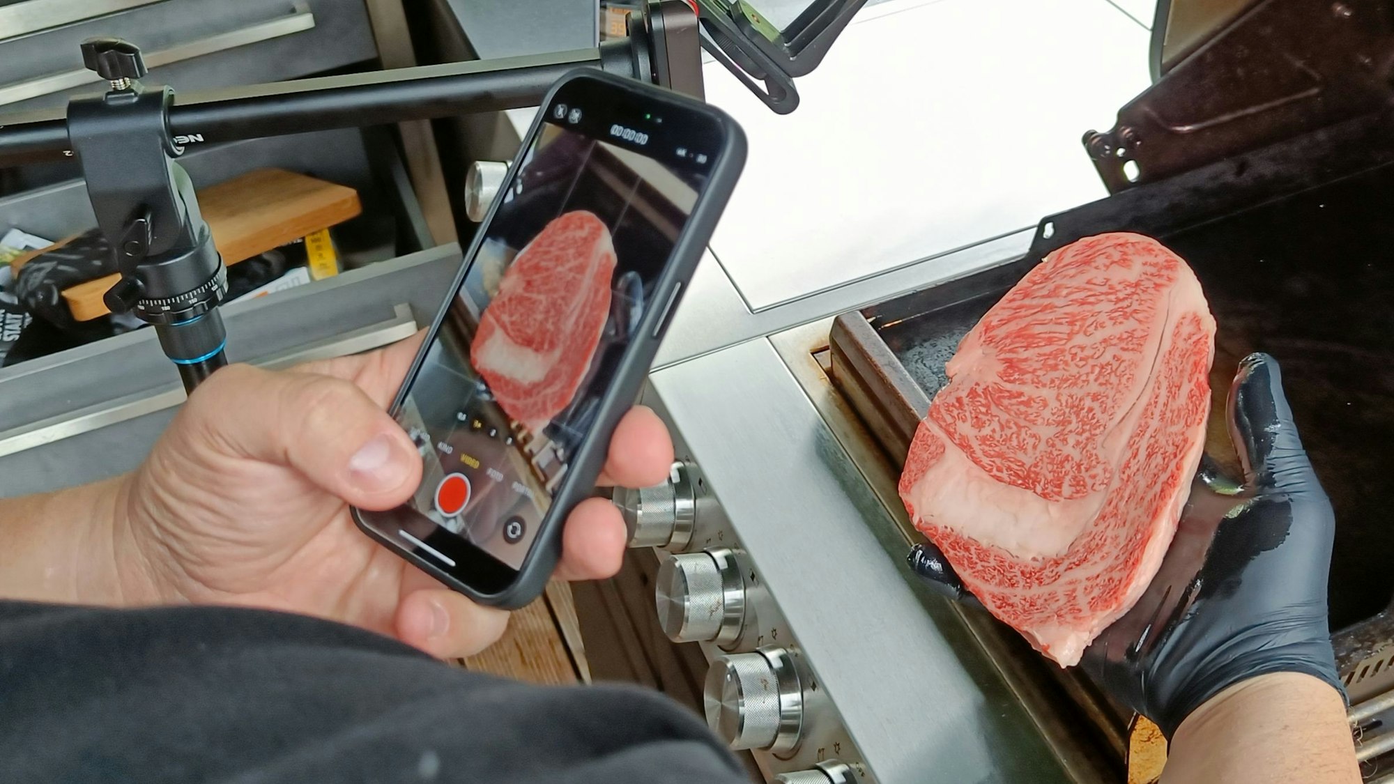 Steak in der Hand, Handy drauf gerichtet, vor einem Grill.