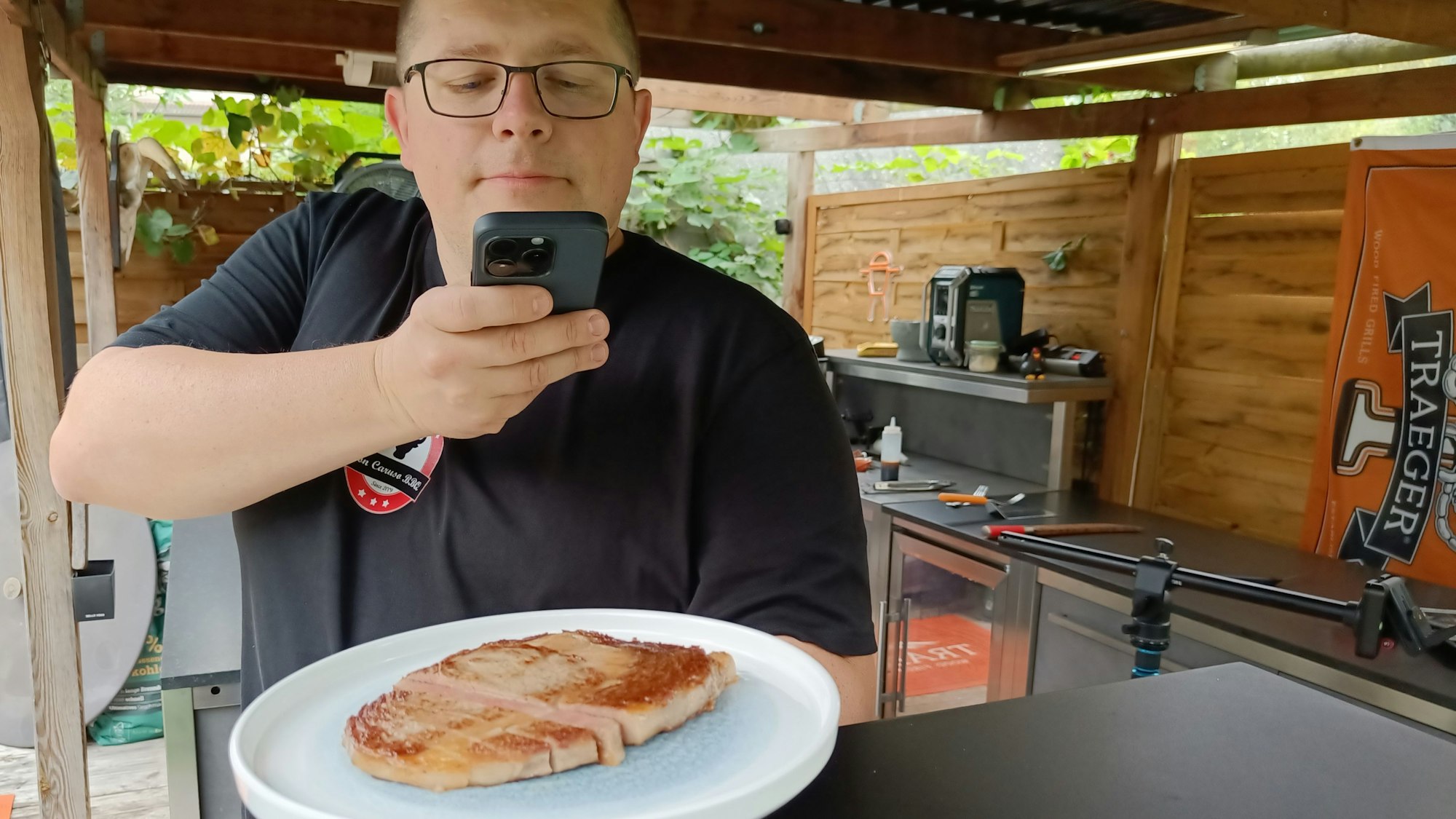 Ein Steak in der Hand, das Handy drauf gerichtet. Vor einem Grill.