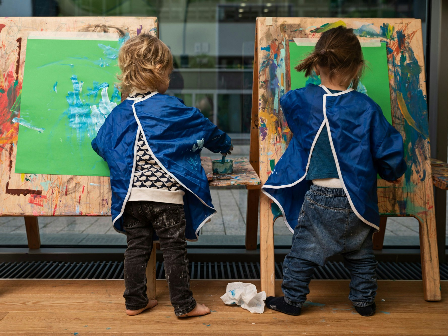 Zwei Kinder malen Bilder an einer Staffelei. Sie tragen Malkittel und sind von hinten zu sehen.