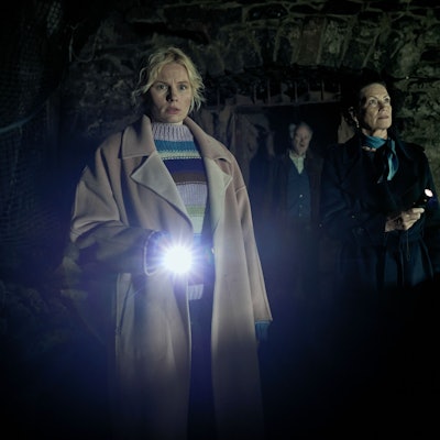 Drei Personen laufen durch ein dunkles Gemäuer. Eine blonde Frau leuchtet den Weg mit der Taschenlampe aus.