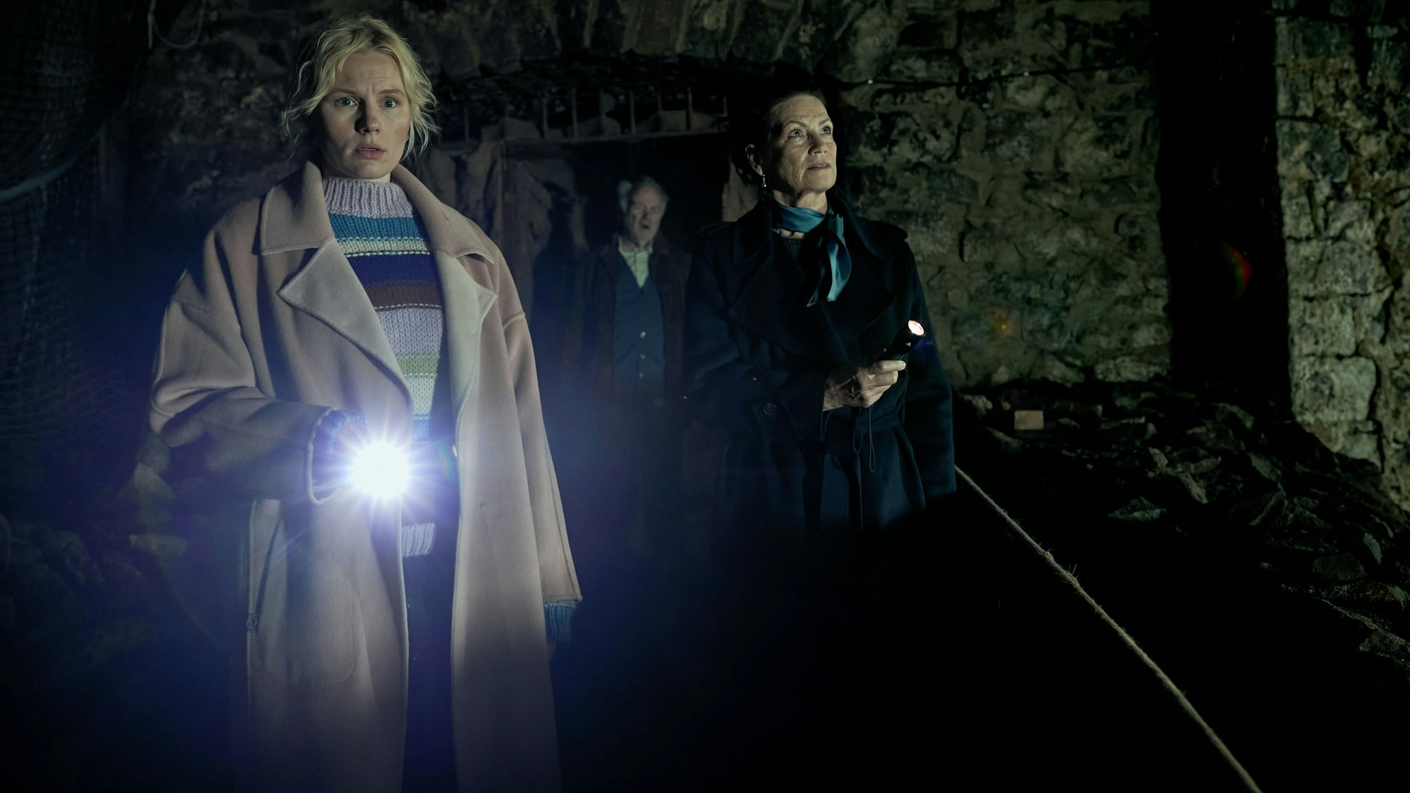 Drei Personen laufen durch ein dunkles Gemäuer. Eine blonde Frau leuchtet den Weg mit der Taschenlampe aus.
