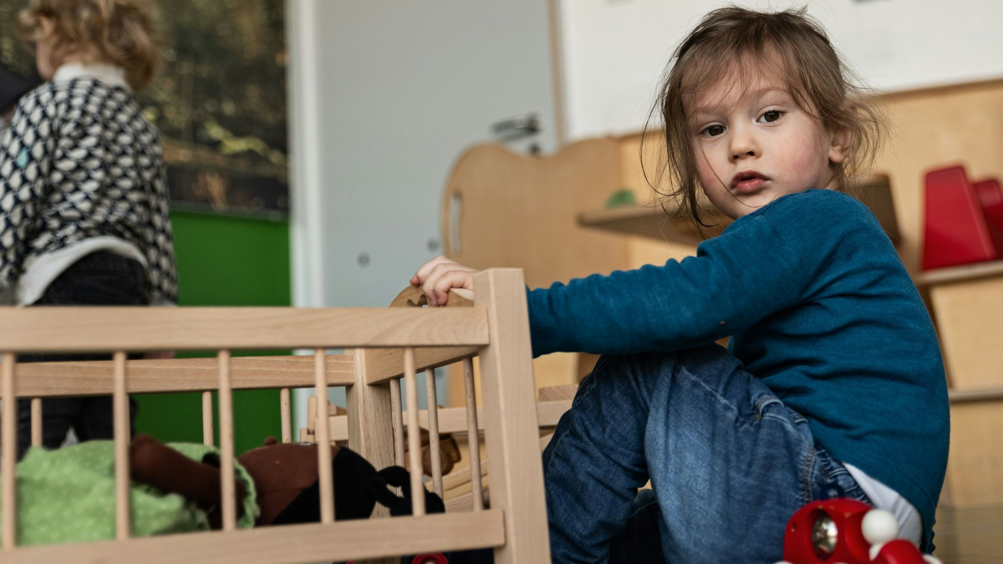 Ein Kind sitzt auf dem Boden neben einem Puppenbett und schaut in die Kamera.
