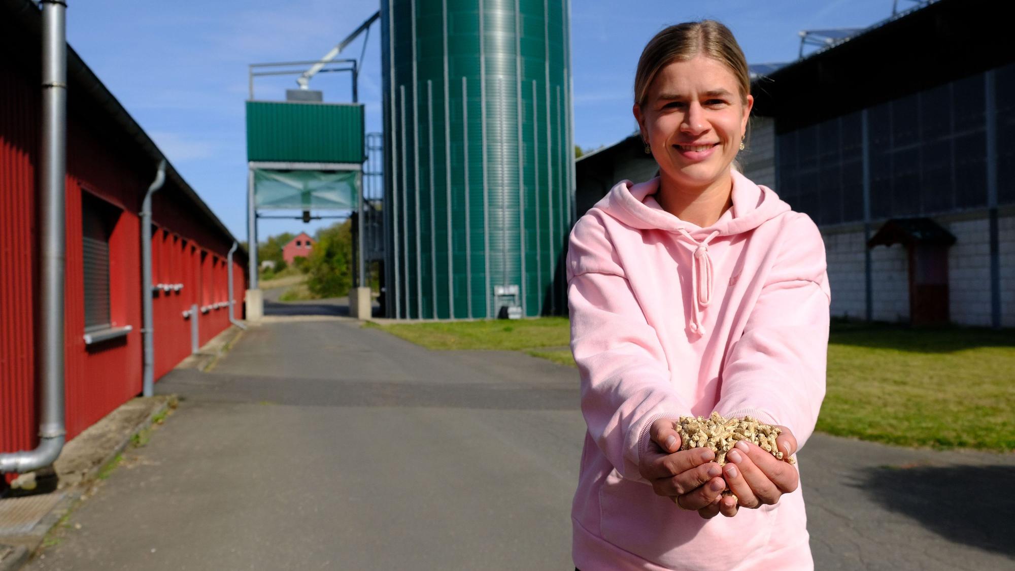 Catharina Hartges hält mit beiden Händen Holzpellets in die Kamera. Im Hintergrund ist ein Siloturm zu erkennen.
