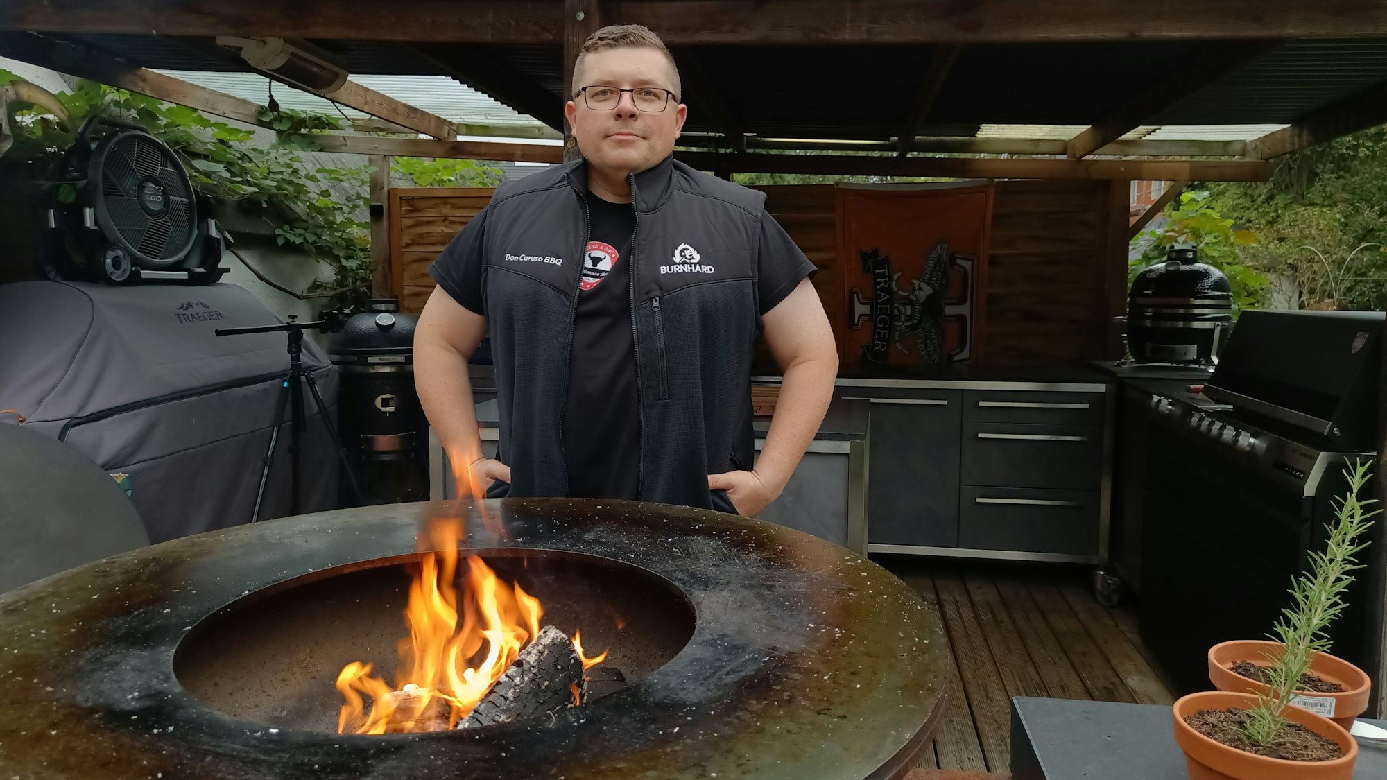 Tomanek steht vor einem Holzdach, darunter verschiedene Grills. Im Vordergrund eine Feuerschale.
