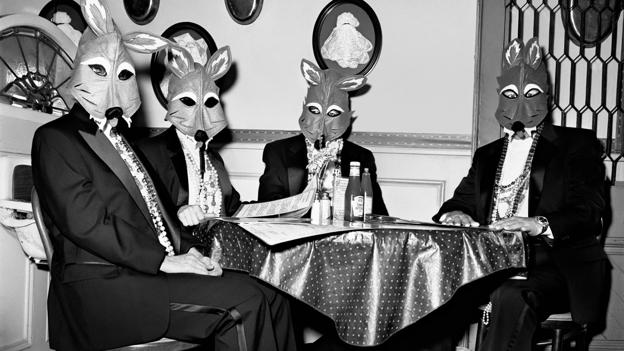 Vier Männer mit Fuchsmasken sitzen in Fracks an einem Tisch.