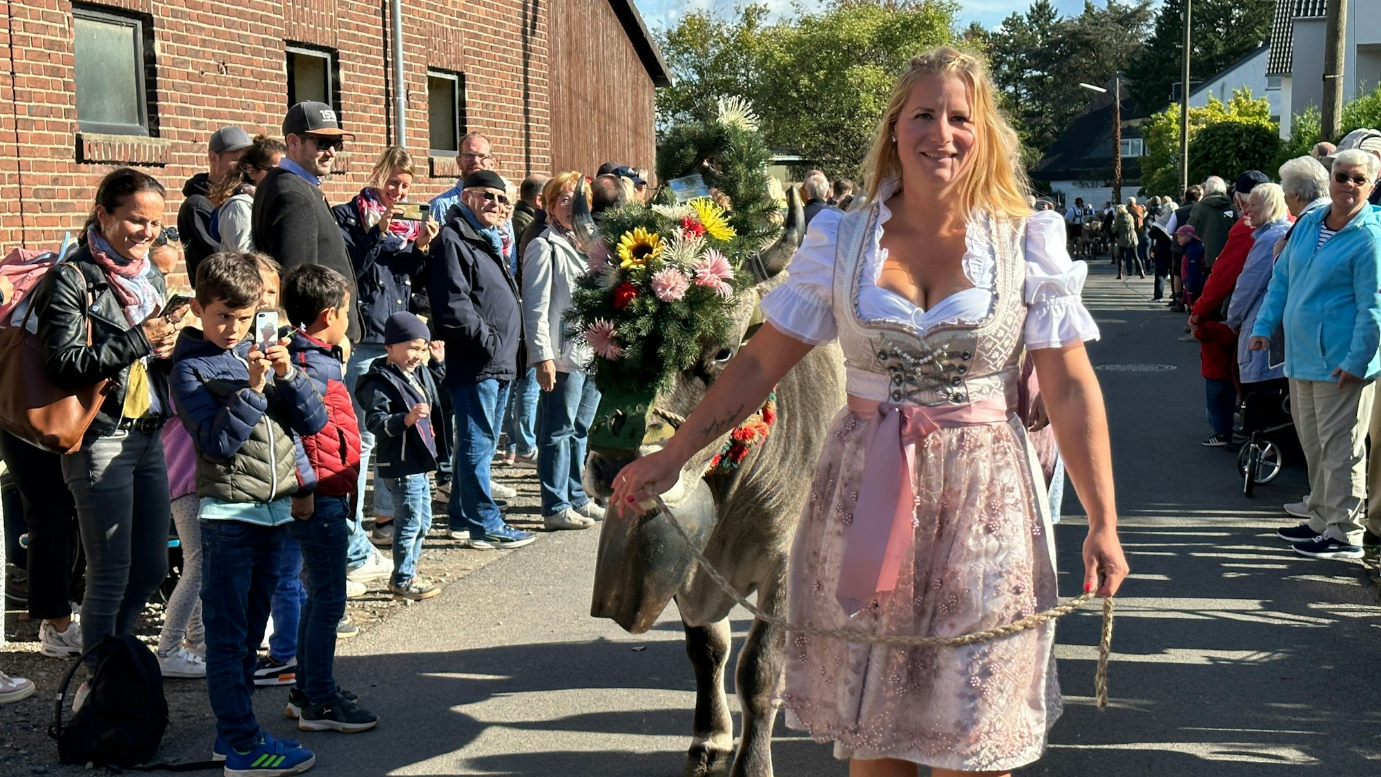 Eine Frau im Dirndl führt eine Kuh mit Kuhglocke durch eine menschengesäumte Straße.
