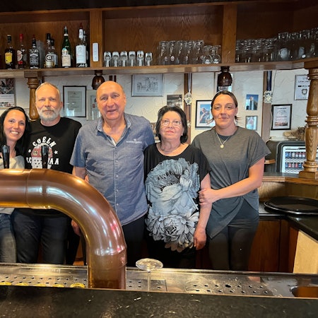 Zwei Männer und drei Frauen stehen hinter einer Theke, im Hintergrund stehen Gläser und Flaschen in einem offenen Schrank.