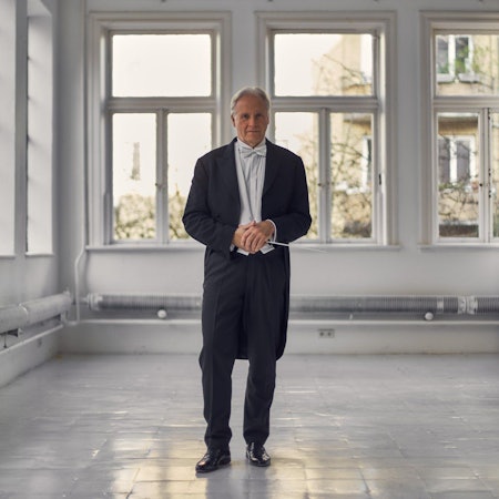 Stenz steht in einem kargen Raum, an dessen Wände einige Rohre sichtbar sind. Er trägt einen Frack, eine weiße Fliege und hält seinen Taktstock.