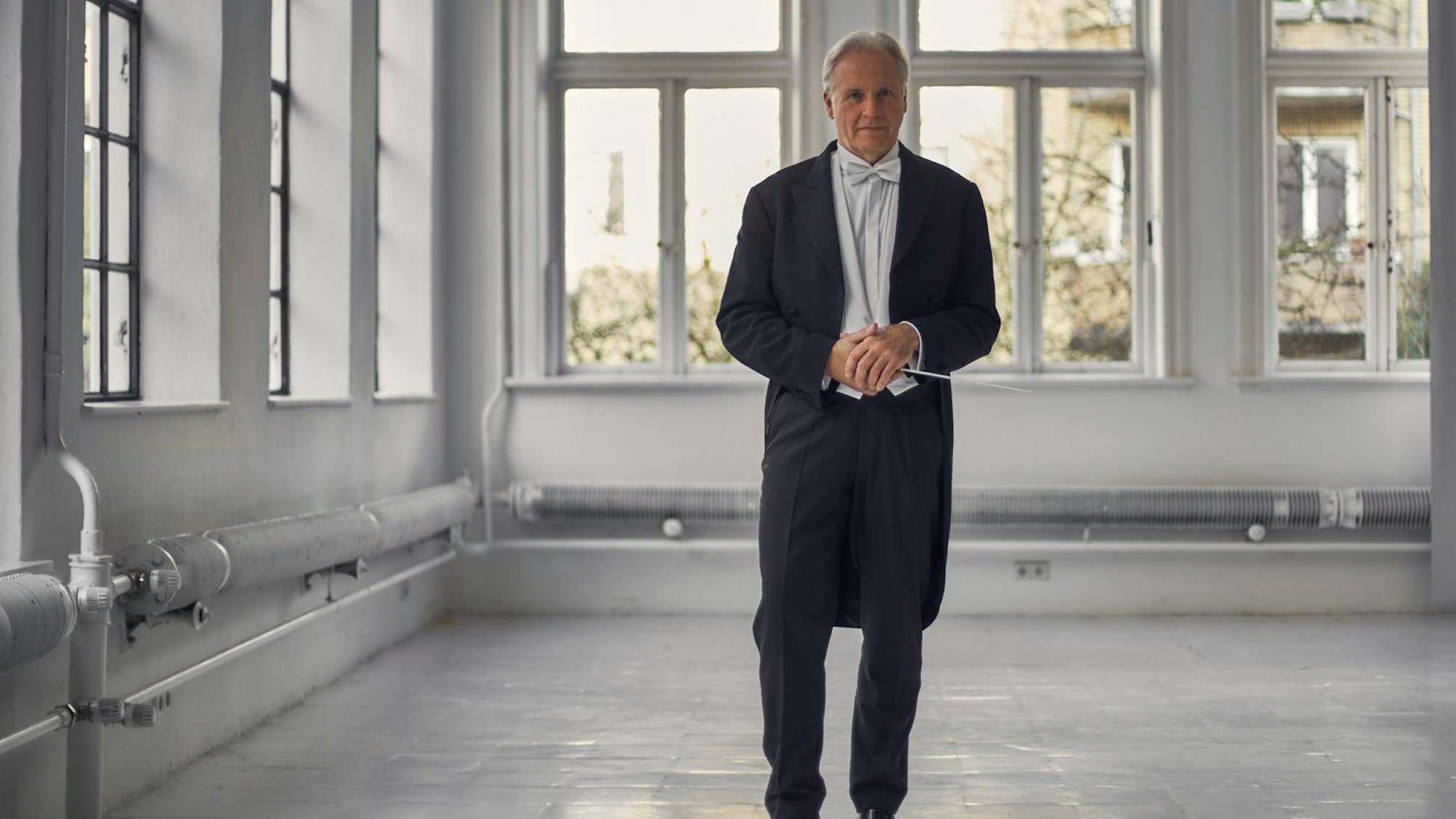 Stenz steht in einem kargen Raum, an dessen Wände einige Rohre sichtbar sind. Er trägt einen Frack, eine weiße Fliege und hält seinen Taktstock.