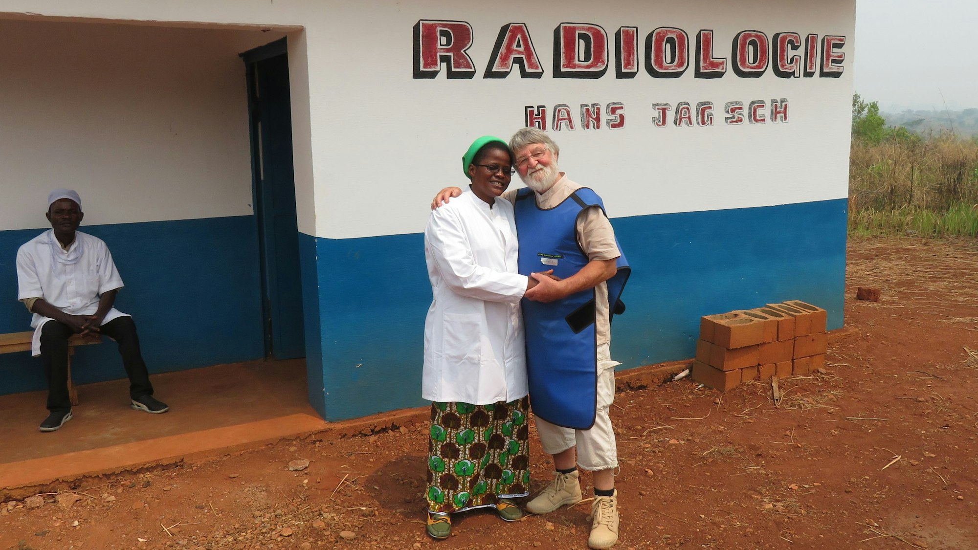 Vor einem blau und weiß gestrichenem einfachen Haus stehen Hans Jagsch und eine Ordensschwester. An der Hauswand steht in roten Buchstaben „Radiologie Hans Jagsch“.