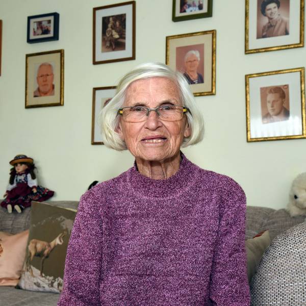 Waltraud Geisen sitzt in ihrem Wohnzimmer auf dem Sofa und schaut in die Kamera, hinter sich an der Wand sind Familienbilder zu sehen.