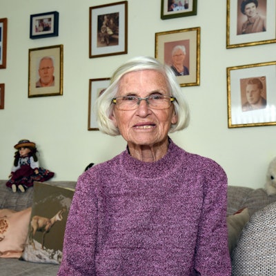 Waltraud Geisen sitzt in ihrem Wohnzimmer auf dem Sofa und schaut in die Kamera, hinter sich an der Wand sind Familienbilder zu sehen.