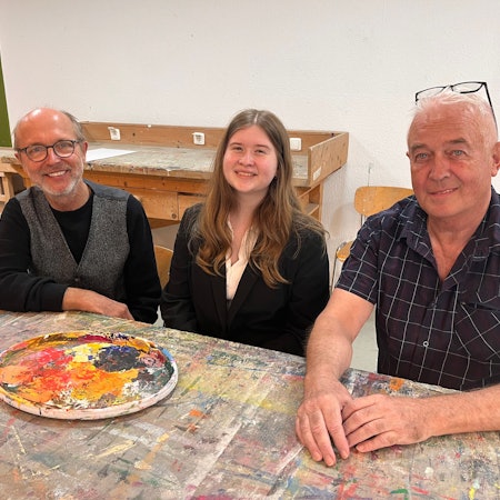 Zwei Männer und eine Frau sitzen an einem Tisch, auf dem Malfarben in einem runden Teller stehen.